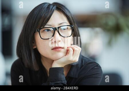Cloe up head and épaules portrait shot of attactive jeune Asian femme portant des lunettes stylées reposant menton sur sa main regardant loin Banque D'Images