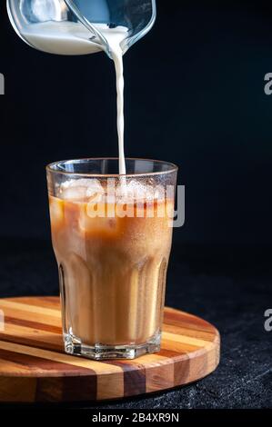 Le Lait Est Versé Dans Du Café Glacé Dans Le Grand Verre Sur Fond Sombre. Boisson D'Été Rafraîchissante Concept. Banque D'Images