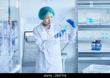 Tir horizontal moyen long de jeune ouvrier de laboratoire asiatique portant un manteau blanc, des gants de protection et un bouchon tenant le tube à essai avec du liquide bleu Banque D'Images