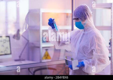 Pharmacienne féminine non reconnaissable portant un uniforme de protection jetable en choisissant un tube à essai, vue latérale horizontale, prise moyenne Banque D'Images