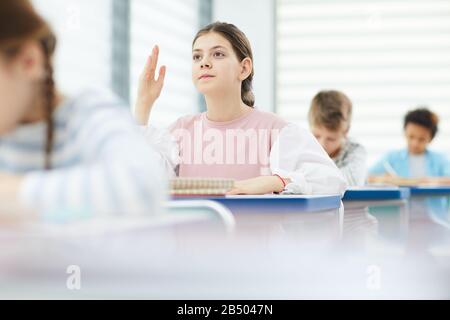 Portrait horizontal d'une jeune fille active et intelligente de douze ans assise à la réception de l'école désireuse de répondre soulevant sa main en classe, espace de copie Banque D'Images