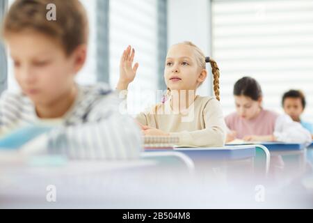 Portrait horizontal de la jeune fille pensive assise à la réception de l'école levant la main et posant des questions à l'enseignant, copier l'espace Banque D'Images