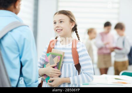Portrait moyen horizontal d'une jeune fille joyeuse de onze ans avec sac à dos tenant des manuels discutant avec son ami de l'école pendant la pause, espace de copie Banque D'Images