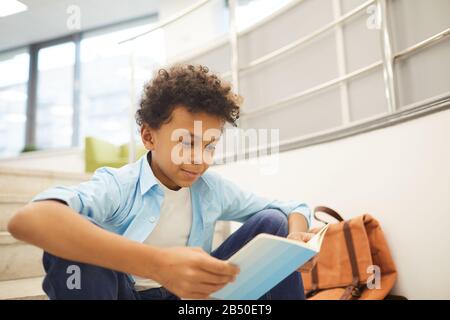 Portrait moyen horizontal d'un curieux garçon africain américain lecture livre intéressant assis seul sur les escaliers, espace de copie Banque D'Images