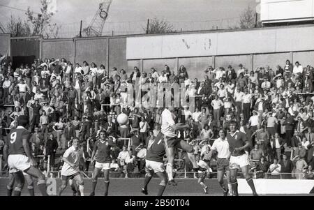 Années 1970, historique, fans regardant un match de football debout sur les terrasses, à Millwall, FC, South London, Angleterre, ROYAUME-UNI. Les terrasses de football sont un certain nombre de marches en béton avec des barrières métalliques installées à de nombreux endroits pour empêcher les gens de descendre la pente. Banque D'Images