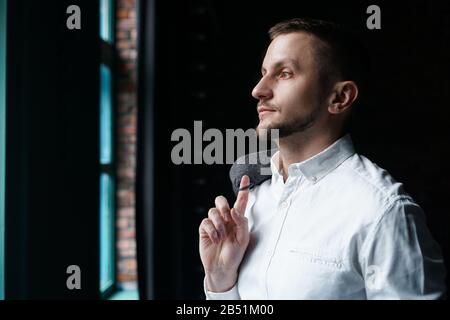 Vue en profil d'un jeune homme d'affaires, habillé d'une chemise blanche debout près de la fenêtre sur un mur sombre et regardant la fenêtre. Banque D'Images