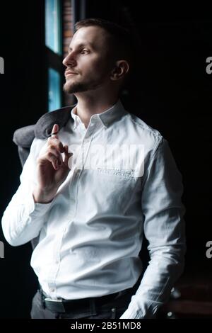 Vue en profil d'un jeune homme d'affaires, habillé d'une chemise blanche debout près de la fenêtre sur un mur sombre et regardant la fenêtre. Photo verticale Banque D'Images