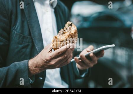 Gros plan d'un jeune colar blanc mangeant des aliments indésirables tout en travaillant sur un smartphone à l'extérieur Banque D'Images