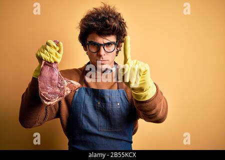 Un jeune boucher qui tient des steaks debout sur fond jaune isolé Pointant avec doigt vers le haut et expression en colère, ne montrant aucun gestur Banque D'Images