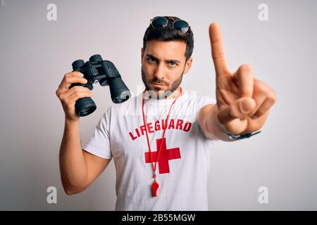 Jeune homme de sauveteur avec barbe portant un t-shirt avec croix rouge et lunettes de soleil en utilisant le sifflement Pointant avec doigt vers le haut et expression en colère, montrant aucun ge Banque D'Images