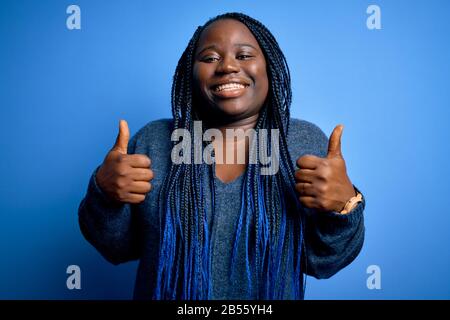 Afro-américaine plus femme de taille avec des tresses portant un pull décontracté sur fond bleu signe de succès faire un geste positif avec la main, pouce vers le haut malle Banque D'Images