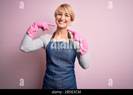 Jeune femme blonde plus propre avec des cheveux courts portant un tablier et des gants sur fond rose souriant montrant et pointant avec les doigts dents et Banque D'Images