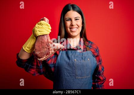 La jeune boucherie, qui tient de la viande de boeuf fraîche et brute, se pit sur fond rouge avec un visage heureux debout et souriant avec un sourire confiant montrant les dents Banque D'Images