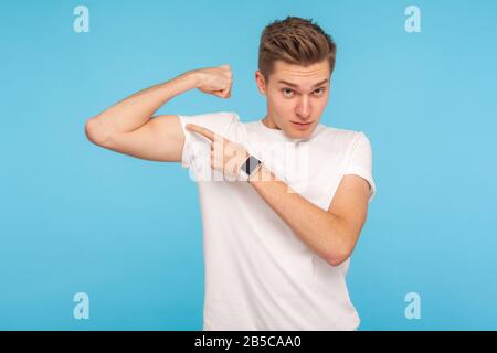 Regardez ma force ! Portrait d'un jeune homme mince dans un t-shirt blanc pointant des biceps, montrant le muscle sur la main levée et le sentiment de pouvoir, confiant dans le corps. Banque D'Images