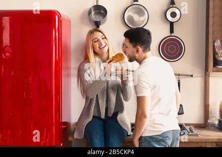 Petit déjeuner amusant. Jeune femme nourrissant son mari avec croissant dans la cuisine Banque D'Images