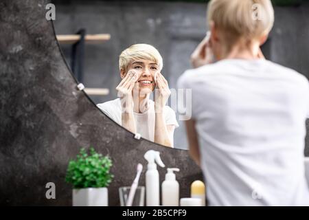 Femme Souriante Appliquant Une Lotion Hydratante Debout Dans La Salle De Bains Intérieur Banque D'Images