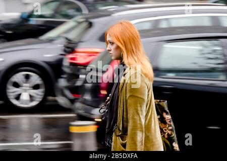 Belgrade, Serbie - 24 septembre 2019 : jeune femme jeune à tête rouge marchant seule sur la rue de la ville pluvieuse dans le flou de mouvement, et des voitures mobiles derrière lui Banque D'Images