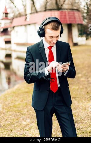 jeune gars dans un costume avec une cravate rouge écoutant de la musique dans de grands écouteurs dans la nature Banque D'Images