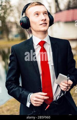 jeune homme d'affaires dans un casque. le gars écoute de la musique sur la nature dans le parc. homme dans un costume noir et cravate rouge Banque D'Images