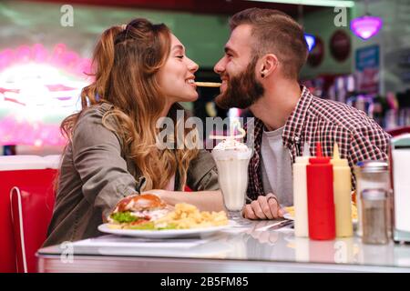 Image d'un drôle de jeune couple aimant s'asseoir dans un café rétro lumineux manger des frites à l'intérieur. Banque D'Images