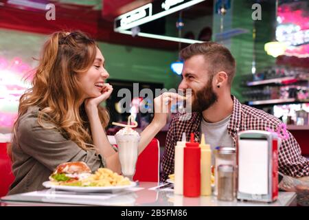 Image d'un jeune couple souriant et positif s'asseoir dans un café rétro lumineux manger des frites à l'intérieur. Banque D'Images