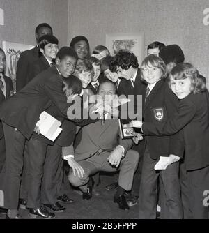 1972, historique, 'Take it on the Chin'.... légende britannique de la boxe Henry Cooper s'amusant avec un groupe d'écoliers en uniforme lors d'une visite à une école secondaire du centre-ville dans le sud de Londres, Angleterre, Royaume-Uni. Banque D'Images