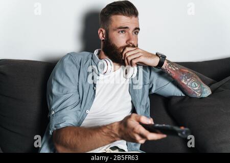 Bel homme ennuyé et barbu se reposant sur un canapé à la maison, regardant la télévision Banque D'Images