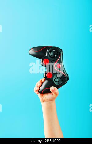 La main d'un enfant tient triomphalement la manette sur un fond bleu. Concept créatif minimaliste de la jeune génération de joueurs. Gagner en ligne Banque D'Images