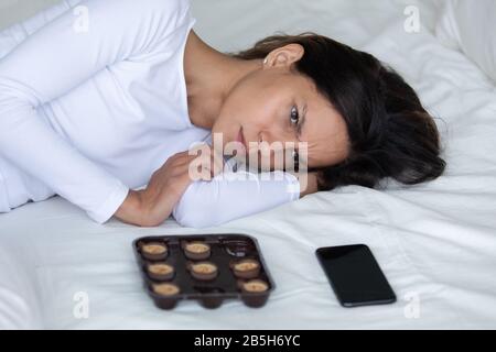 Fille déprimée posée dans le lit avec des bonbons et smartphone Banque D'Images