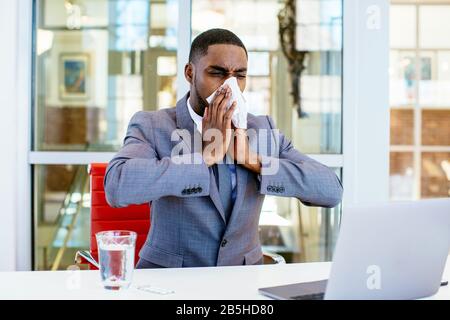 Portrait d'un jeune homme malade en costume d'affaires soufflant son nez tout en étant assis derrière le bureau au travail avec l'ordinateur Banque D'Images