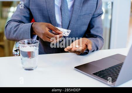 Détail des mains des hommes en costume d'affaires et prendre une pilule médicale avec de l'eau tout en étant assis derrière le bureau au travail avec l'ordinateur Banque D'Images