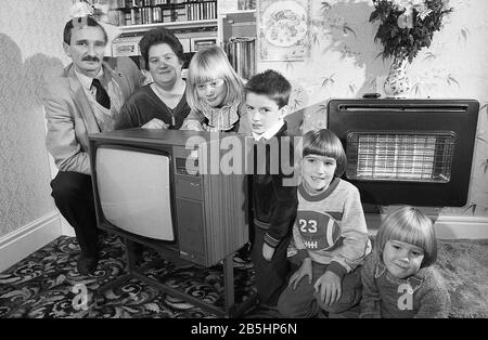 Années 1980, historique, à l'intérieur d'une salle avant, une famille, mère, père et leurs quatre jeunes enfants, alignés pour une photo, accroupissant à côté de leur nouvelle télévision assis sur un support en métal à roues, Angleterre, Royaume-Uni. Un feu de barre électrique mural de l'époque vue sur la photo. Banque D'Images