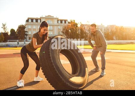 Homme et femme faisant crossfit à l'extérieur. Une fille sportive pousse un pneu dans le stade au coucher du soleil. Sportifs. Banque D'Images