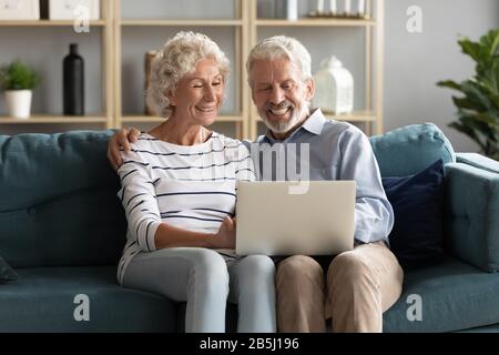 Un mari et une femme souriants et âgés se reposent sur un canapé avec ordinateur portable Banque D'Images