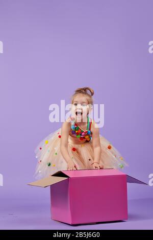 Une petite fille drôle enfant dans une magnifique robe de vacances regarder à l'intérieur d'une grande boîte de couleur cadeau, sur un fond violet isolé. Surprise d'anniversaire Banque D'Images