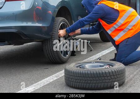 Un homme dans un gilet de sécurité orange change un pneu plat sur une route. Gros plan les mains à la roue d'une voiture cassée. Remplacement d'une roue à l'aide d'un skrewdriver Banque D'Images