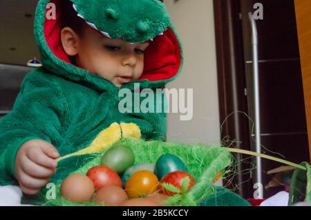 Mignon petit enfant dans le costume vert de dinosaure jouant avec les oeufs de pâques colorés.. Banque D'Images