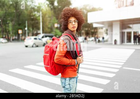 Un jeune homme afro-américain avec un sac à dos rouge traverse la route à un passage pour piétons dans la ville. Un étudiant est en attente de traverser la route. Concep Banque D'Images