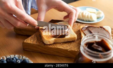 Étaler le beurre de noix de chocolat sur du pain grillé. Les mains féminines frottez le chocolat sur le pain sandwich. Préparation du déjeuner ou du petit déjeuner Banque D'Images