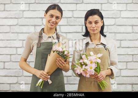 Taille vers le haut portrait de deux fleuristes femmes souriant à l'appareil photo tout en tenant de beaux bouquets de printemps tout en posant contre le mur de briques Banque D'Images