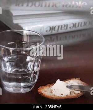 En arrière-plan, des livres de cuisine, au premier plan un verre d'eau, une petite tranche de pain traditionnel avec du beurre ou du fromage. Banque D'Images