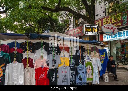 Guilin, Chine - 9 mai 2010 : rue commerçante piétonne de Zhengyang. Le vendeur de vêtements ambulants affiche sa marchandise sous une tente sous le feuillage vert. Banque D'Images