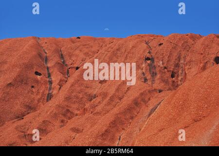 Territoire du Nord, Australie centrale, une demi-lune se lève sur les crêtes et les ondulations de la roche de grès rouge solide d'Uluru contre un ciel bleu clair Banque D'Images