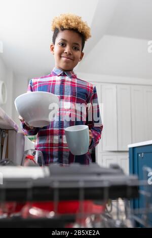 Portrait De Boy Aider Avec Les Corvées À La Maison Par Stacking Vaisselle Dans Le Lave-Vaisselle Banque D'Images