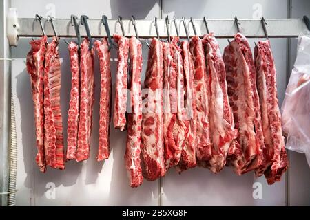 Rangée de côtes de porc fraîches et brutes accrochées au réfrigérateur sur des crochets métalliques dans la boucherie ou la cuisine du restaurant Banque D'Images