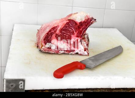 Un morceau frais de viande de jambon à l'œil de côtes brutes sur la planche à découper à côté du gros couteau rouge Banque D'Images