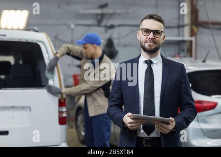 Portrait moyen horizontal de jeune homme d'affaires caucasien debout avec tablette PC regardant l'appareil photo, spécialiste du service automobile travaillant derrière lui Banque D'Images
