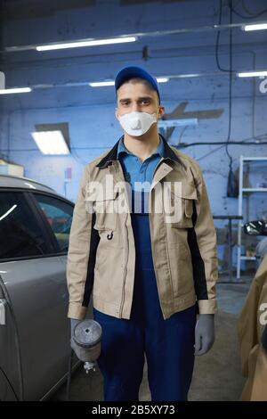 Portrait vertical moyen long tourné d'un peintre de voiture professionnel portant un vêtement de travail de protection et un masque sur le visage regardant l'appareil photo Banque D'Images