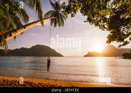 Silhouette d'hommes swing avec coucher de soleil sur l'île tropicale en arrière-plan. Baie d'El Nido. Philippines. Banque D'Images