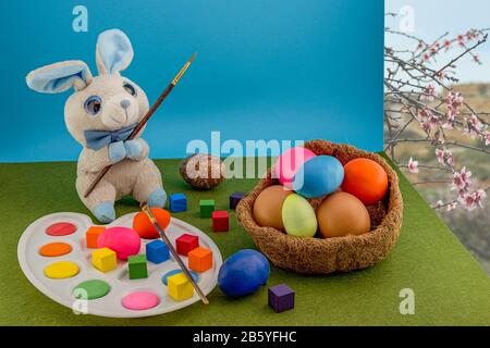 Oeufs de pâques colorés dans le nid de paille. Un lapin doux gris avec une palette et une brosse peint des œufs de Pâques. Cubes multicolores autour. L'herbe verte CA Banque D'Images